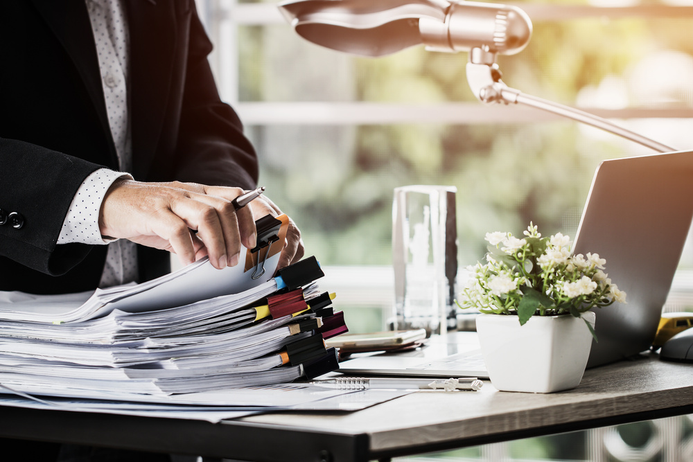 Businessman hands holding pen for working in Stacks of paper files searching information business report papers and piles of unfinished documents achieves on laptop computer desk in modern office